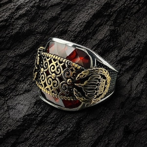 Könnte beinhalten: Ein silberner Ring mit einem großen, facettierten, roten Edelstein. Der Ring hat aufwendige goldene Filigranarbeiten und ein goldenes Emblem an der Seite. Der Ring ist vor einem dunklen, strukturierten Hintergrund platziert.
