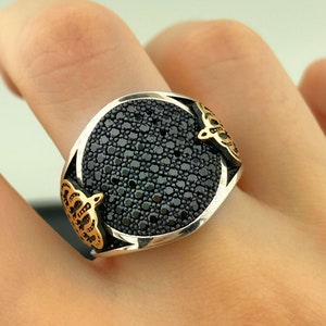 May include: A silver ring with a large, round black stone set with numerous small black gems. The ring has gold-colored decorative accents on each side. The ring is shown on a finger.