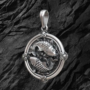 Puede incluir: Colgante de plata con dos peces koi dentro de un marco circular. El colgante presenta detalles intrincados, incluyendo escamas y motivos de plantas acu&aacute;ticas. La argolla es un simple lazo para una cadena.