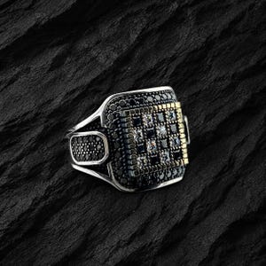 Könnte beinhalten: Ein silberner Ring mit quadratischer Fassung, besetzt mit schwarzen und klaren Steinen. Der Ring hat ein geometrisches Design mit einem goldfarbenen Akzent auf einer Seite. Das Band hat eine abgerundete Form mit einem rechteckigen schwarzen Stein auf jeder Seite.
