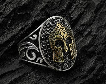 May include: Silver ring with an oval face featuring a gold-colored helmet design. The helmet is set against a black stone background, surrounded by black stones. The ring has intricate silver detailing on the band.