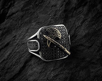 May include: A silver ring with a black stone setting, adorned with a gold-colored axe and sword design. The ring features a rectangular face covered in small black stones, with additional black stones on the band.