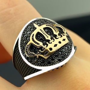 May include: A silver ring with a gold crown design. The crown is centered on a black stone setting. The ring has a thick band and a detailed crown design, showcasing a regal aesthetic.