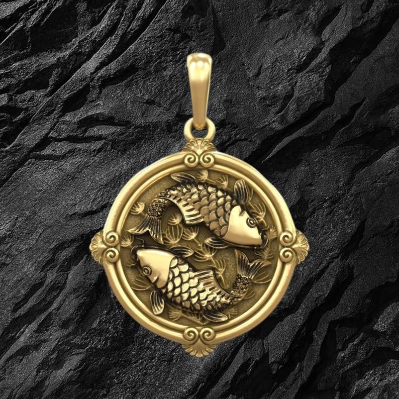 Puede incluir: Colgante dorado con dos peces en un dise&ntilde;o circular, representando el signo zodiacal de Piscis. El colgante tiene detalles intrincados y un borde decorativo, con un lazo en la parte superior para una cadena.