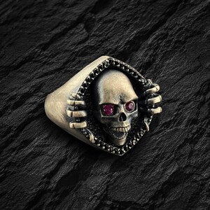 May include: A silver skull ring with ruby-colored eyes and black accents. The skull is framed within a diamond-shaped setting, with the ring's band featuring a textured design. The ring is set against a dark, textured background.