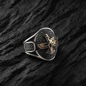 May include: Silver ring with a black stone oval centre featuring a gold-coloured bird emblem. The ring has a rectangular black stone accent on the side. The ring is set against a dark, textured background.