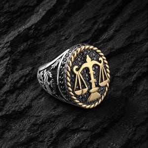 May include: Silver ring with a gold-colored scale of justice emblem. The ring features a black stone border and a rope-like gold border around the emblem. The ring's band has a floral design.