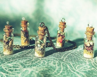 Könnte beinhalten: Fünf kleine Glasflaschen mit Korkstopfen und silbernen Ketten, gefüllt mit verschiedenen bunten Steinen und Kristallen. Die Flaschen sind auf einem grünen Stoffhintergrund angeordnet.