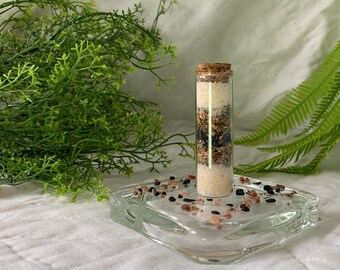 May include: A small glass bottle with a cork stopper filled with layers of dried herbs, crystals, and white salt. The bottle is sitting on a clear glass dish with small pieces of black and pink crystals.