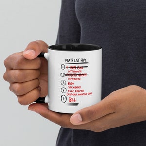 Puede incluir: Taza de cerámica blanca con borde y asa negros. La taza tiene una lista manuscrita en negro que dice "Death List Five" con cinco nombres y descripciones. Los nombres están tachados en rojo.