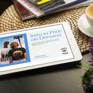 Può includere: Un tablet bianco mostra il testo "Intro to Print-on-Demand" con un'immagine di due persone che indossano magliette. Sono visibili anche una tazza di caffè, riviste e una piccola pianta viola.