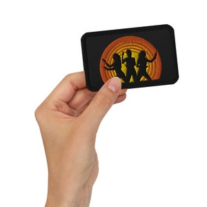 May include: Black embroidered patch featuring a silhouette of three figures in front of a yellow and orange concentric circle design. The patch is rectangular with a black border and is held in a hand.
