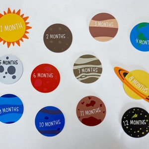May include: A set of 12 colorful, round, felt circles with a white outline and white text. Each circle represents a month of the year, with the text "1 Month" through "12 Months". The circles are arranged in a circle around a yellow sun with orange rays.