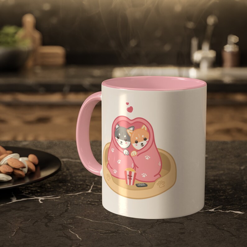 May include: White ceramic mug with a pink rim and handle. The mug features a cartoon illustration of two dogs cuddling under a pink blanket with hearts and a bowl of popcorn. The text "woof" is printed on the bottom of the mug.