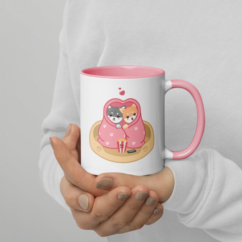 May include: A pink and white ceramic mug with a design of two dogs cuddling under a blanket with popcorn and a remote control. The mug has a pink rim and handle.