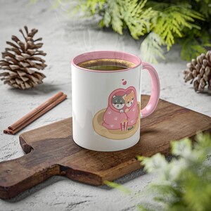 May include: A white ceramic mug with a pink rim and handle. The mug features a cartoon illustration of two dogs snuggled under a pink blanket with a bucket of popcorn. The dogs are looking at each other and there are hearts above them.