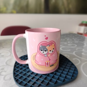 May include: A pink ceramic mug with a design of two cartoon dogs cuddling under a blanket. The dogs are a gray and white husky and a brown and white Shiba Inu. The mug is on a blue and gray square-shaped coaster.