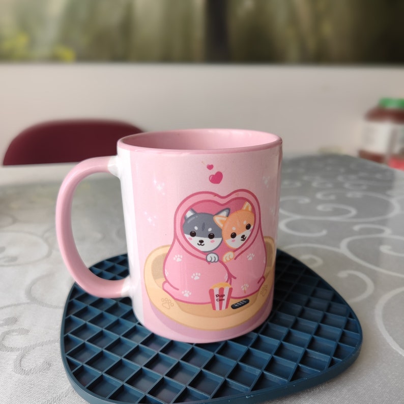 May include: A pink ceramic mug with a design of two cartoon dogs cuddling under a blanket. The dogs are a gray and white husky and a brown and white Shiba Inu. The mug is on a blue and gray square-shaped coaster.