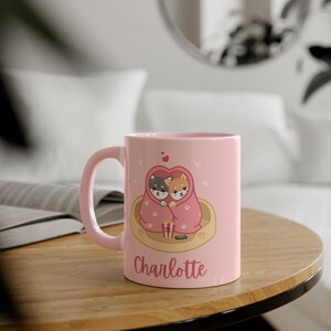 May include: Pink ceramic mug with a design of two dogs cuddling under a blanket with popcorn and a remote control. The mug has the name "Charlotte" printed on the bottom.
