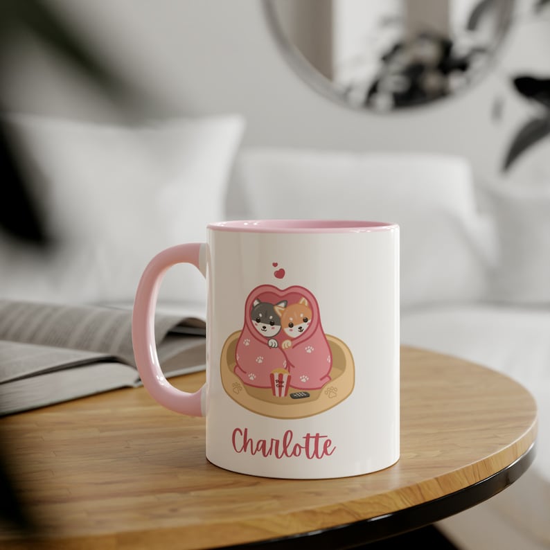 May include: A pink and white ceramic mug with a pink rim. The mug features a cartoon illustration of two dogs cuddling under a blanket with a heart shape. The text "Charlotte" is printed on the mug.