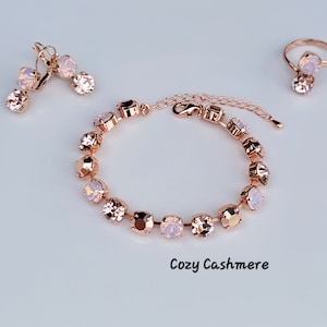 May include: A rose gold jewelry set featuring a bracelet, earrings, and a ring. The bracelet and earrings have a series of light pink gemstones. The ring has a single gemstone. The text "Cozy Cashmere" is at the bottom.