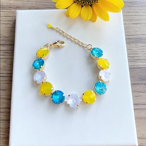 May include: A gold-toned bracelet featuring a series of colorful, round gemstones. The gemstones are in shades of yellow, blue, and white. The bracelet is displayed on a white surface, with a yellow sunflower in the background.