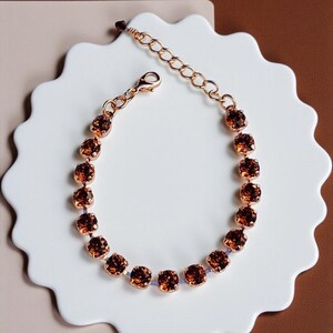 Könnte beinhalten: Ein roségoldenes Armband mit runden, bernsteinfarbenen Kristallen. Das Armband hat eine zarte Kette und einen Karabinerverschluss. Die Kristalle sind gleichmäßig verteilt und erzeugen einen funkelnden Effekt. Das Armband wird auf einer weißen Oberfläche mit gewelltem Rand präsentiert.