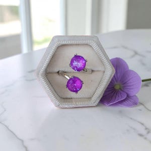 May include: A silver-toned bypass ring featuring two round, vibrant purple faceted stones set in a hexagonal, light-colored velvet ring box. The ring is open-ended, with each stone held by four prongs, showcasing a bright, reflective, and polished metallic band.