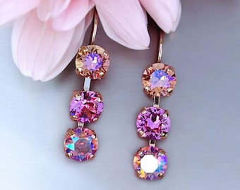 May include: A pair of dangle earrings with three iridescent pink and peach-colored crystals. The earrings have a gold-colored metal setting and are displayed near a soft pink flower.