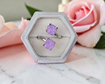 May include: A silver-toned open ring featuring two round, textured, light purple druzy stones set in prong settings. The ring is displayed inside a hexagonal, light gray velvet ring box with a single slot for the jewelry piece.