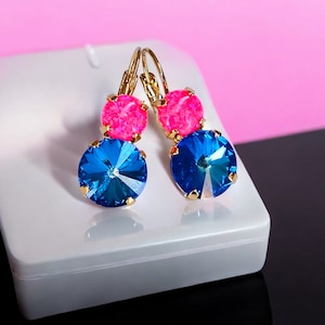 May include: A pair of gold earrings with blue and pink crystal stones. The earrings have a dangling design with a larger blue crystal stone at the bottom and a smaller pink crystal stone above it.