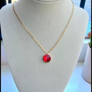 May include: A gold-colored necklace with a bright red, round pendant. The pendant is a faceted gemstone set in a gold-colored bezel. The necklace is displayed on a white mannequin.