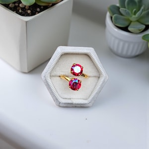 May include: Two gold rings with large, round, red gemstones are displayed in a gray velvet ring box. The rings are set against a white background, with small potted plants in the background.