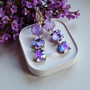 May include: Dangle earrings with a gold-tone metal setting. Each earring has three purple, round crystals, the bottom crystal of each with an iridescent effect. The earrings are displayed on a white surface, with purple flowers in the background.