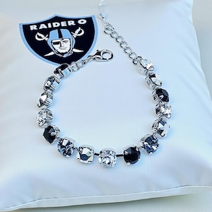 Op de afbeelding: Een zilverkleurige armband met afwisselend zwarte en heldere vierkante kristallen. De armband wordt weergegeven op een wit kussen naast een Raiders-logo.