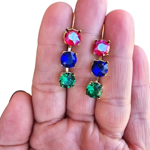 May include: A pair of gold-toned drop earrings, each featuring three colorful, round gemstones. The gemstones are arranged vertically, with a red-pink top stone, a blue middle stone, and a green bottom stone. The earrings are held up by a gold hook.