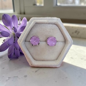 May include: A pair of round stud earrings featuring textured, light purple druzy stones set in silver-toned metal. The earrings are displayed in a hexagonal, cream-colored velvet jewelry box, positioned next to a purple flower blossom.