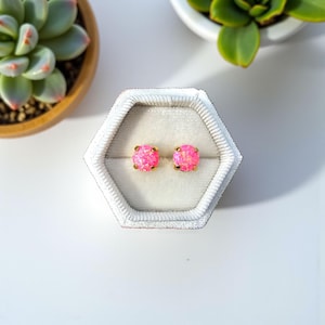 Könnte beinhalten: Ein Paar rosa Opal-Ohrstecker mit goldfarbenen Stiften, präsentiert in einer weißen sechseckigen Schmuckschatulle. Die Ohrringe haben eine quadratische Form und eine schimmernde, hellrosa Farbe. Der Hintergrund umfasst Topf-Sukkulenten.