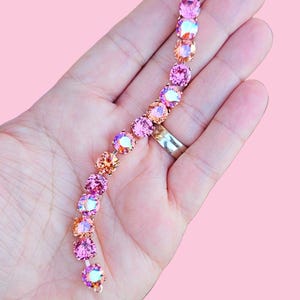 Op de afbeelding: Een delicate armband met een gradi&euml;nt van roze en oranje iriserende kristallen. De armband is gemaakt van een gouden metalen ketting en heeft een sluiting.
