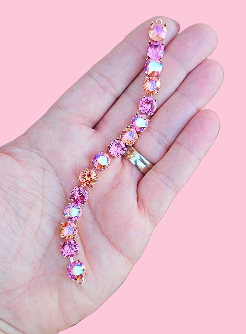 Op de afbeelding: Een delicate armband met een gradi&euml;nt van roze en oranje iriserende kristallen. De armband is gemaakt van een gouden metalen ketting en heeft een sluiting.