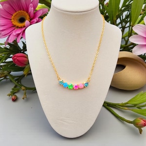 Puede incluir: Un collar dorado con una serie de piedras preciosas de colores. Las piedras están dispuestas en una línea curva, con piedras azules, verdes y rosas. El collar se exhibe en un maniquí blanco, sobre un fondo de flores rosas.