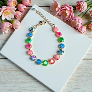 Peut inclure: Un bracelet doré orné de pierres précieuses rondes et colorées dans les tons rose, vert, bleu et irisé. Le bracelet est présenté sur une surface blanche, avec des fleurs roses en arrière-plan. Le bracelet a une fermeture à fermoir homard.