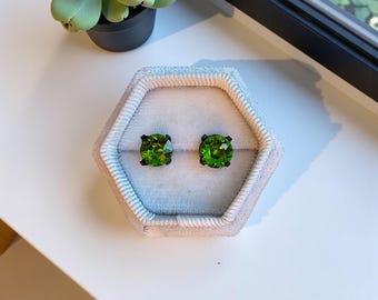 May include: A pair of green gemstone stud earrings displayed in a light gray velvet jewelry box. The earrings have a square cut and are set in a dark metal. A small succulent plant is visible in the background.