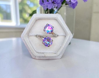 May include: A silver bypass ring featuring two round, iridescent, multi-colored stones that display shifting hues of pink, blue, and purple. The ring is displayed in a hexagonal, light-colored velvet ring box with a central slot for the band.