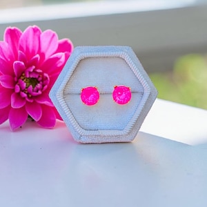 Könnte beinhalten: Ein Paar leuchtend pinkfarbene Ohrstecker mit goldenen Stiften, präsentiert in einer hellblauen, sechseckigen Schmuckschatulle. Im Hintergrund ist eine leuchtend pinkfarbene Dahlie zu sehen, die der Komposition Farbe verleiht.