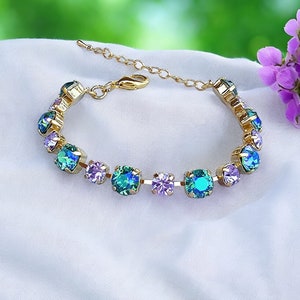 Może przedstawiać: Złota bransoletka z łańcuszka z zapięciem na zatrzask. Bransoletka jest ozdobiona naprzemiennie jasnofioletowymi i turkusowymi okrągłymi kryształami.