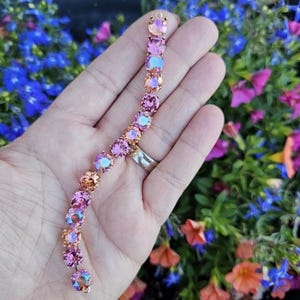 Op de afbeelding: Een armband gemaakt van roze en oranje iriserende kristallen. De armband is op een hand met een ring aan de middelvinger.