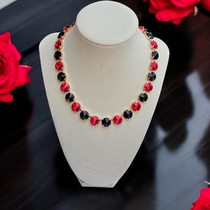 May include: A gold-toned necklace with alternating red and black round gemstones. The necklace is displayed on a white bust, with red roses in the background. The gemstones are evenly spaced and create a striking contrast.