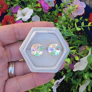 May include: A pair of iridescent crystal stud earrings displayed in a light gray hexagonal jewelry box. The earrings have a gold-colored post and a round, faceted crystal that reflects multiple colors. The background features colorful flowers and greenery.
