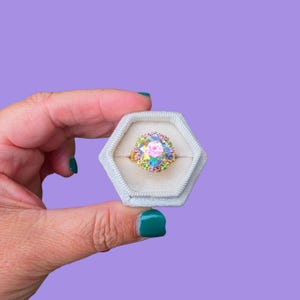 May include: A gold ring with a large, iridescent, multi-colored gemstone center, surrounded by smaller, colorful stones. The ring is set in a light gray, hexagonal ring box against a purple background.